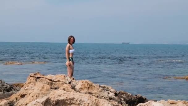 Caldo attraente bruna donna in bikini e rosa occhiali da sole in piedi sulla spiaggia rocciosa in riva al mare. Ritratto di giovane donna sulla spiaggia — Video Stock