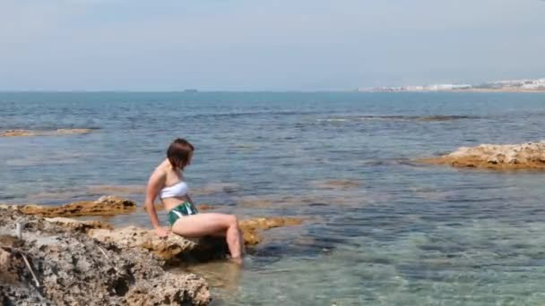 Charmig varm kvinna med vacker kropp i vit och grön Bikini sittande på sten på stranden. Attraktiv kvinna doppa benen i havet — Stockvideo