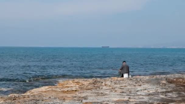 Elderly lonely man sitting on empty seashore with a bait-casting rod and fishing. Lonesome isolated fisherman on beach — Stock Video