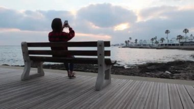 Genç kadının silueti bankta oturup deniz ve gün batımının fotoğraflarını çekiyor. Gezinti fotoğraflarını çeken kadın silueti