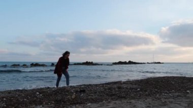 Kot pantolon ve sıcak kırmızı gömlek boş kayalık plajda duran ve gün batımında suya taş atma sevimli yalnız kadın