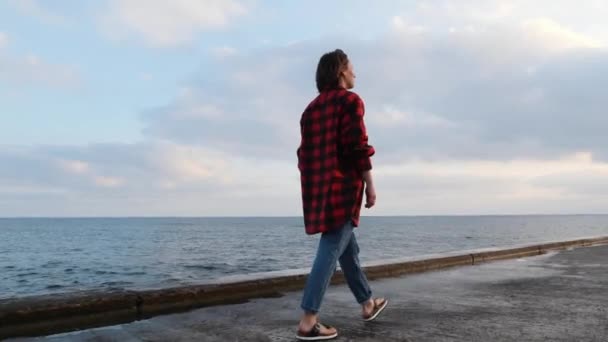 Aantrekkelijke jonge brunette vrouw wandelen langs de kust bij zonsondergang. Schattig charmant meisje in casual slijtage wandelen op de promenade door de zee — Stockvideo