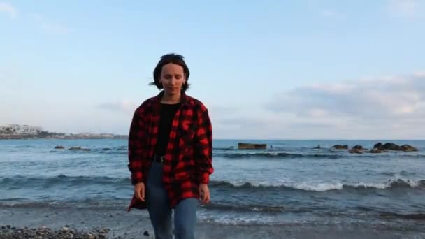 Giovane donna bruna caucasica in camicia a quadri calda rossa e jeans che camminano sulla spiaggia vuota alla sera ventosa — Video Stock
