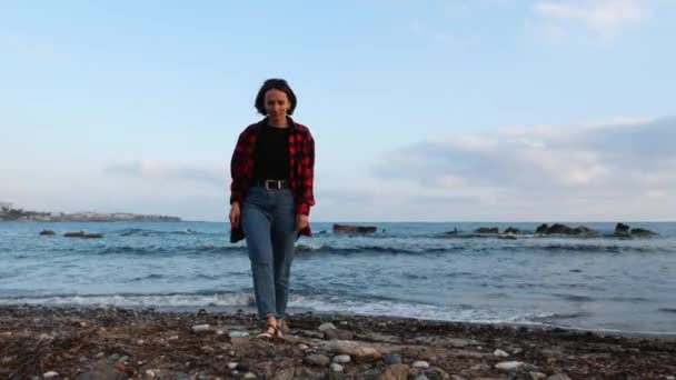 Aantrekkelijke eenzame vrouw wandelen langs de kust bij zonsondergang en kijken naar de camera. Jong brunette meisje wandelen op het strand door de zee — Stockvideo