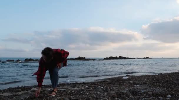 Меланхоличная молодая женщина в тёплой красной рубашке и джинсах собирает камни на скалистом пляже и бросает их в тёмное море вечером — стоковое видео