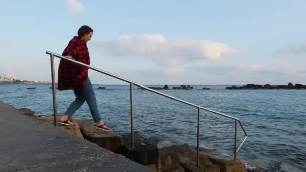 Mulher morena jovem desce ao mar ao longo de passos de concreto segurando para corrimão. Mulher atraente descendo para o mar — Vídeo de Stock