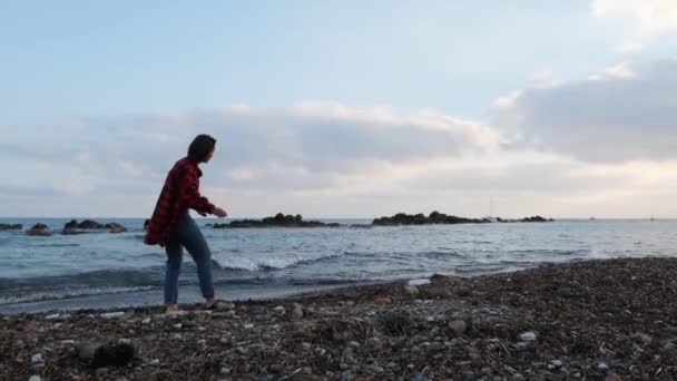 Mujer solitaria y melancólica en ropa casual caliente arroja piedras al agua al atardecer con el mar y el cielo nublado en el fondo — Vídeo de stock