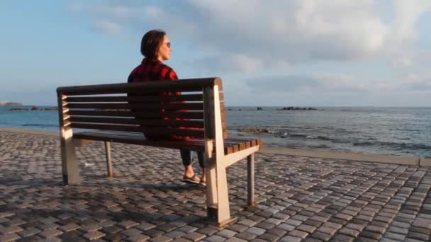 Einsame traurige Frau in warmem kariertem Hemd und Jeans, die auf einer Holzbank am leeren Kai sitzt und den Sonnenuntergang betrachtet — Stockvideo