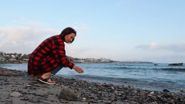 Mujer solitaria hermosa con camisa roja cálida y jeans sentados en la costa rocosa vacía, recogiendo y examinando piedras — Vídeos de Stock