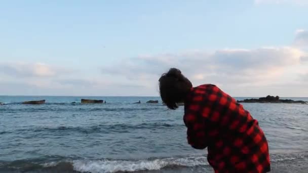年轻的有吸引力的黑发女人在红色温暖的衬衫扔小石头到海在空的岩石海滩，特写后视图 — 图库视频影像