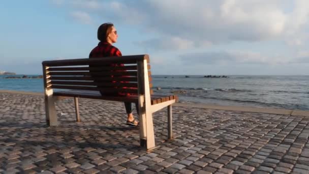Kaukasische junge Frau im warmen karierten Hemd sitzt auf einer Holzbank an der leeren Promenade und blickt abends aufs Meer — Stockvideo