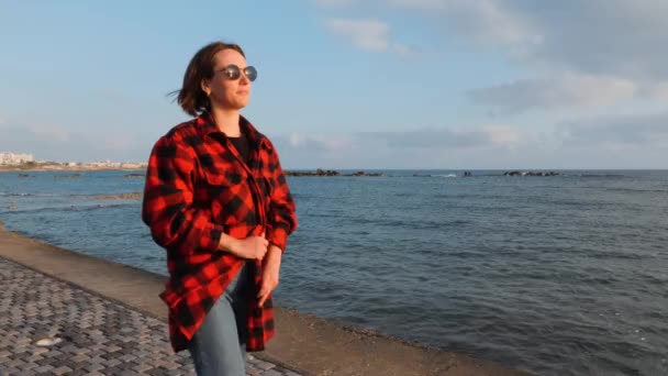 Carino bruna femmina in camicia a quadri rossa e jeans che camminano lungo la riva del mare la sera. Ragazza che cammina sul lungomare vuoto — Video Stock