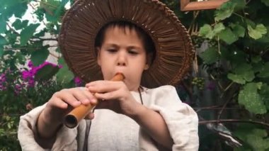 Yeşil bahçede ahşap müzik aleti üzerinde çalan hasır şapkalı küçük sevimli çocuğun yakından görünümü. Mutlu çocuk flüt çalar