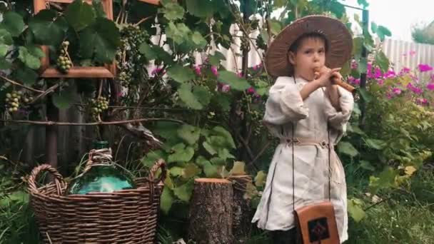 Bájos aranyos kaukázusi gyerek a falusi ruházat és kalap szórakozik a faluban, és játszik a fuvola szőlő a háttérben — Stock videók