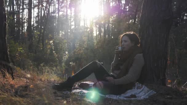 Happy charming woman drinking coffee sitting on plaid outside. Beautiful brunette female in cozy sweater and warm scarf nostalgic and relaxing in fall forest at sunrise — Stock Video