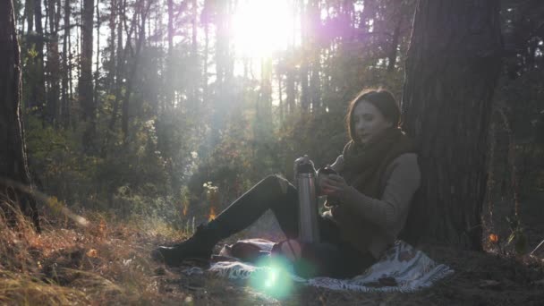 Молодая брюнетка кавказка в теплом свитере и шарфе сидит на клетке в осеннем лесу и наливает и пьет чай из термоса — стоковое видео