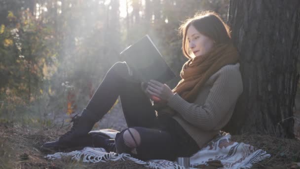 Young good-looking woman in warm cozy clothes reading book, holding cup of tea or coffee and drinking in autumn forest at sunrise — Stock Video