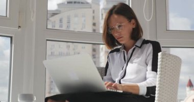 Katı giyinmiş ve gözlüklü kızgın bir iş kadını modern ofiste dizüstü bilgisayarda çalışıyor, hızlı bir şekilde e-posta yazıyor, monitöre bakıyor ve üzülüyor.