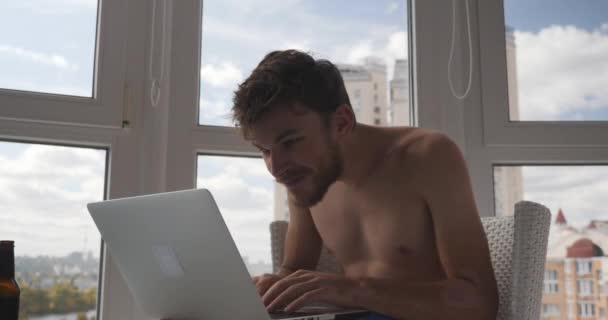 Молодий кавказький чоловічий трейдер в трусах, сидячи на балконі з ноутбуком, виграв угоду на фінансовому ринку, веселий, щасливий і радісний. Щасливий чоловік виграв онлайн відеоігри на комп'ютері — стокове відео