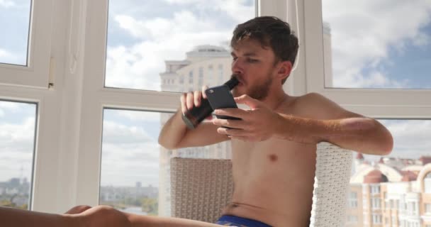 Arrogante patético bêbado jovem caucasiano em cuecas chamando no telefone celular e beber cerveja enquanto sentado na moderna varanda de luxo com grandes janelas — Vídeo de Stock