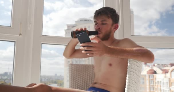술에 취해 속옷 차림의 젊은 남자 가 집에 앉아 휴대 전화로 문자 메시지를 입력하고 술을 마시는 모습 — 비디오