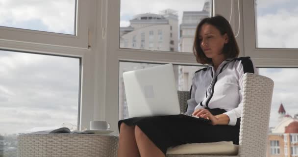 Zmotywowana młoda piękna brunetka biznes kobieta siedzi w nowoczesnym białym krześle, pije kawę i patrzy na duże miasto przez okna biurowe — Wideo stockowe
