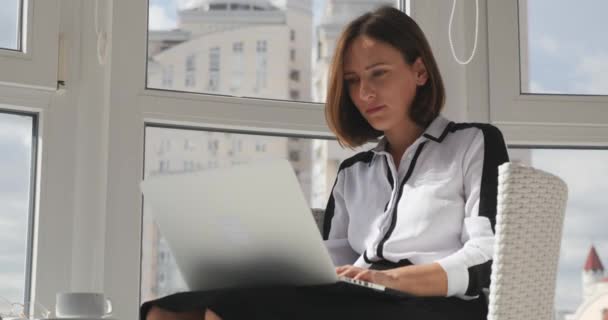 Focado motivado jovem empresário trabalhando em seu escritório com grandes janelas e cidade grande em segundo plano. Convencido funcionário do escritório feminino em roupas rígidas digitando no laptop — Vídeo de Stock