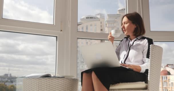 Jeune femme d'affaires élégante confiante travaillant sur ordinateur portable au bureau moderne, buvant du café et regardant la grande ville — Video