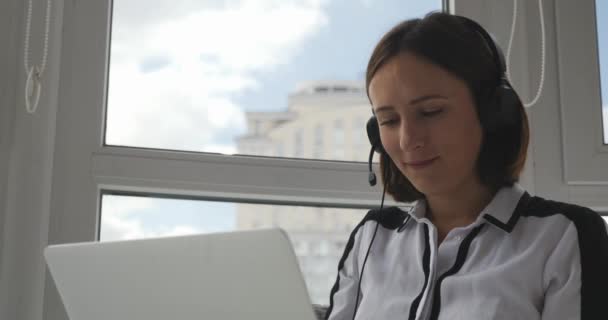 Sonriendo agradable agente de centro de llamadas femenino hablando con los clientes en línea por auriculares. Gerente de soporte femenino en auriculares que consultan a los clientes en el ordenador portátil y chating en línea. Concepto de enseñanza de estudio online — Vídeo de stock