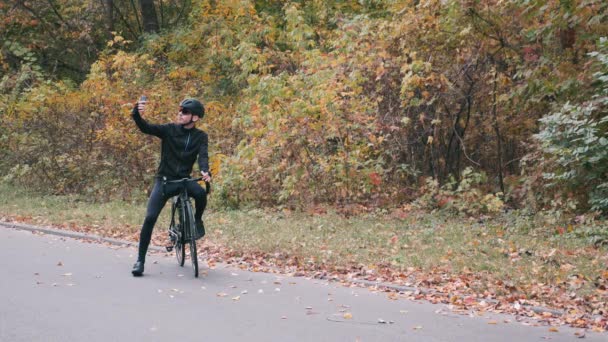 Mladý pohledný profesionální triatlonista v černých cyklistických šatech, černé helmě a brýlích na silničním kole v podzimním lese, který si na telefonu bere selfie. Triatlonový koncept. Mužský cyklista. Zpomalený pohyb — Stock video