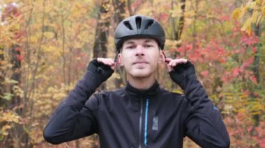 Siyah etkin spor giyimli beyaz bir genç sonbahar parkında siyah bisiklet kaskı ve gözlük takıyor. Erkek bisikletçi bisiklet aksesuarlarını takar