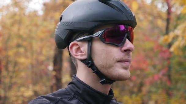 Retrato de un joven guapo triatleta profesional en casco negro y guantes de ciclismo quitándose las gafas deportivas en el parque de otoño. Vista lateral extra cercana — Vídeos de Stock