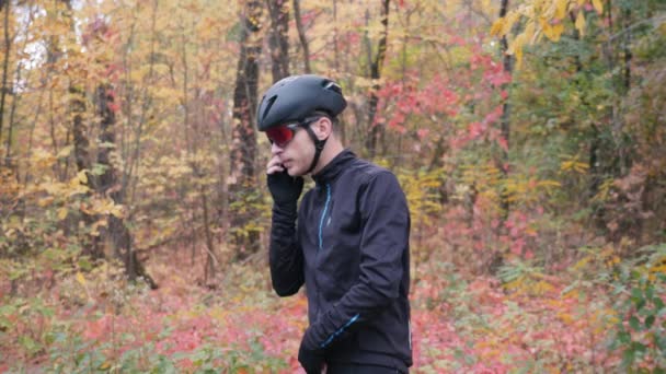 Fiatal profi férfi kerékpáros beszél telefonon edzés előtt a kerékpár őszi parkban. Vonzó ideges kerékpáros srác beszél telefonon őszi erdőben edzés előtt. Lassú mozgás. — Stock videók