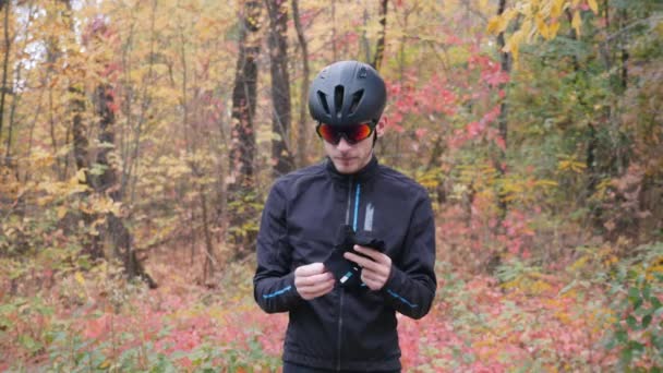 黒のヘルメットと眼鏡のプロの男性サイクリストは、自転車でトレーニングする前にサイクリング手袋に置きます。秋の都市公園で屋外ワークアウトの準備サイクリングマン — ストック動画