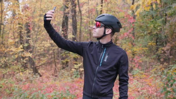 Joven chico deportivo atractivo en ropa de ciclismo tomar selfie en el teléfono inteligente en el parque de la ciudad de otoño. Ciclista profesional tomando fotos de sí mismo en el parque de otoño. Movimiento lento — Vídeo de stock