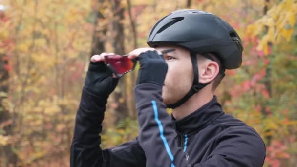 Mladý pohledný profesionální cyklista svléká cyklistické vybavení po tréninku na kole v podzimním parku. Atraktivní kavkazský sportovec sundávající si helmu a brýle po tréninku — Stock video