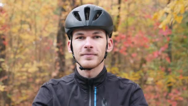 Fiatal profi férfi kerékpáros portréja az őszi parkban. Vonzó motivált sportoló fekete sisakban, aki kerékpáros edzés után leveszi a szemüvegét. Kerékpározás és triatlon koncepció. Lassú mozgás. — Stock videók
