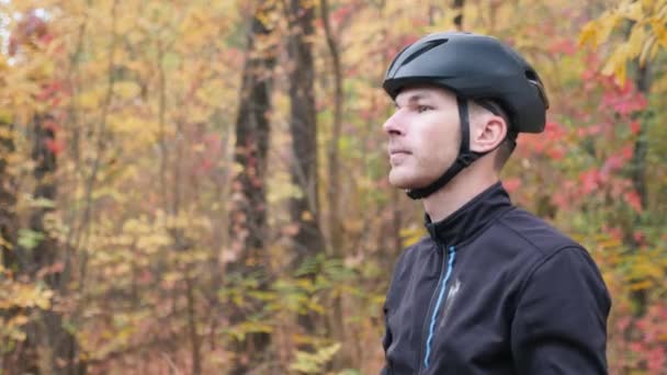 Joven atractivo motivado ciclista masculino quitando equipo de bicicleta. Sonriente atleta profesional guapo se quita el casco de ciclismo negro y gafas después del entrenamiento. Ciclismo hombre en el parque de otoño — Vídeo de stock