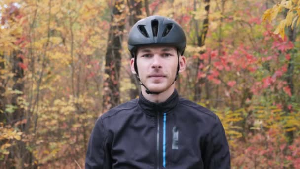 Attractive male cyclist in black helmet and warm thermo sportswear standing in fall city park and puts on sports glasses. Portrait of athlete guy puts on sunglasses in autumn park — Stock Video