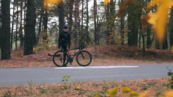 Bisiklet kıyafetleri içinde çekici genç bisikletçi, siyah kask ve güneş gözlüğü takıp sonbahar ormanında poz veriyor. Bisikletli yakışıklı beyaz adam güz parkında eğleniyor. — Stok video