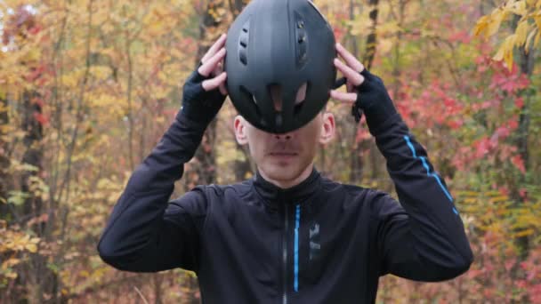 Fiatal férfi sportoló fekete kerékpáros ruhában áll az őszi parkban, és fekete sisakot vesz fel. Kerékpáros koncepció. Férfi biciklizés. Lassú mozgás. — Stock videók