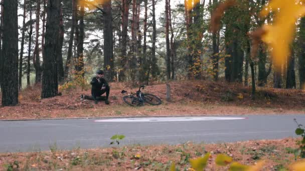 Zelfverzekerde jonge professionele fietser in zwarte helm en zonnebril zittend in het najaarspark en selfie of foto 's nemend via de telefoon. Knappe atleet ontspannen na harde training in het bos — Stockvideo