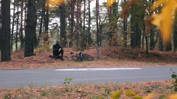 Kasklı ve spor gözlüklü yakışıklı genç erkek sporcu. Sonbahar parkındaki kütüğün üzerinde bisiklet sporu yaptıktan ve dinlendikten sonra oturuyor. Bisikletli adam su içiyor. — Stok video