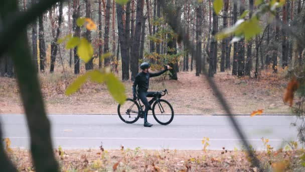 Мотивований молодий професійний велосипедист у чорному шоломі набирає повідомлення, кладе телефон у кишеню і починає їздити на шосейному велосипеді в осінньому парку. Осіннє тренування на велосипеді на свіжому повітрі. Повільний рух — стокове відео