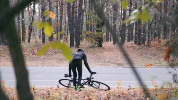 Joven ciclista profesional en ropa de ciclismo y casco negro preparándose para el entrenamiento en bicicleta en el parque de la ciudad de otoño. Atleta deportivo atractivo que comienza a montar en bicicleta de carretera — Vídeo de stock