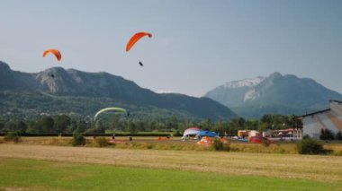 Turuncu yamaç paraşütçüleri Alp Dağları 'ndaki iniş pistine iniyor. Erkek ve kadın insanlar paraşütçüler üzerinde uçuyor.