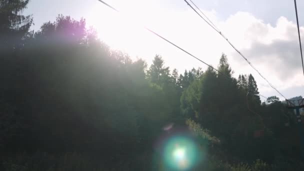 Cables de acero de telesilla entre bosque verde y pinos en montañas contra cielo azul y sol — Vídeo de stock