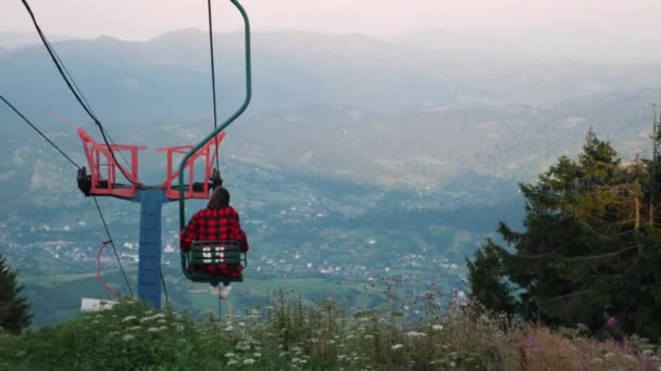 Viajera montada en telesilla con cables de acero desde la cima de la montaña hasta la estación de esquí con paisaje de fondo. Mujer sentada en el asiento de madera del telesilla contra el paisaje de montaña — Vídeos de Stock