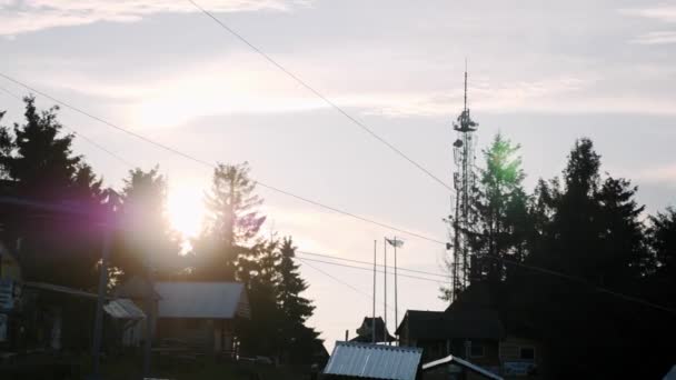 Pine trees on the top of mountain with wood houses and telecommunication station against sun rays and sky. Ski station on the top of hill at sunny summer day — Stock Video