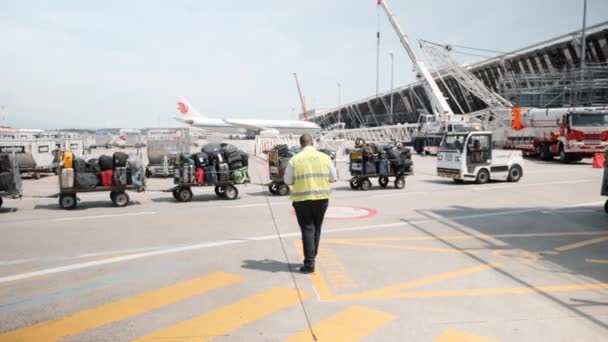 Γενεύη / Ελβετία -Ιούλιος, 22 2019 - μεταφορά αποσκευών στο αεροδρόμιο της Γενεύης. Ειδικό όχημα με βαλίτσες με αεροπλάνο Air China στο παρασκήνιο. Υπάλληλος αεροδρομίου που διαχειρίζεται τη φόρτωση αποσκευών στο αεροπλάνο — Αρχείο Βίντεο